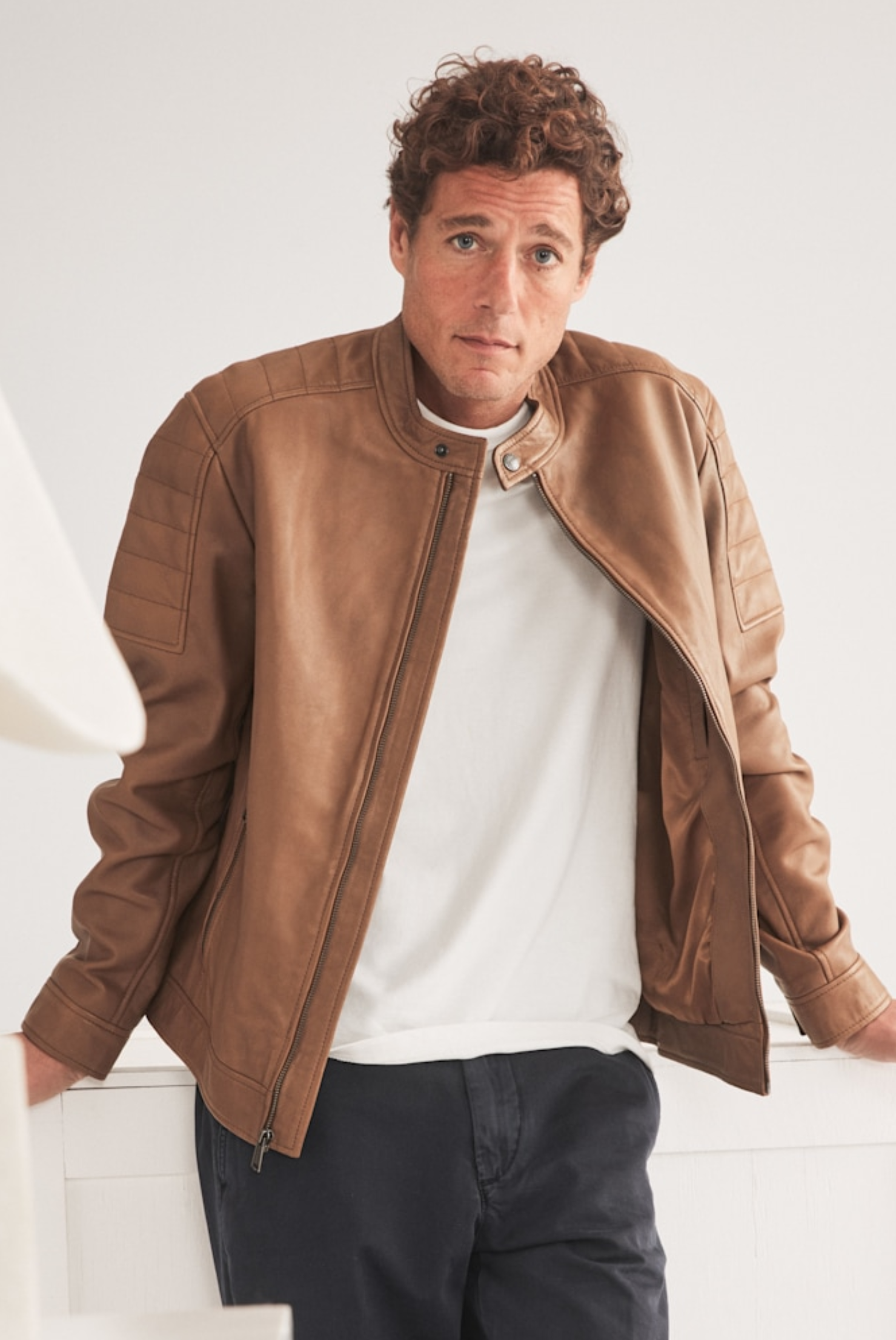 male model standing in a brown leather moto jacket and white t-shirt with dark pants
