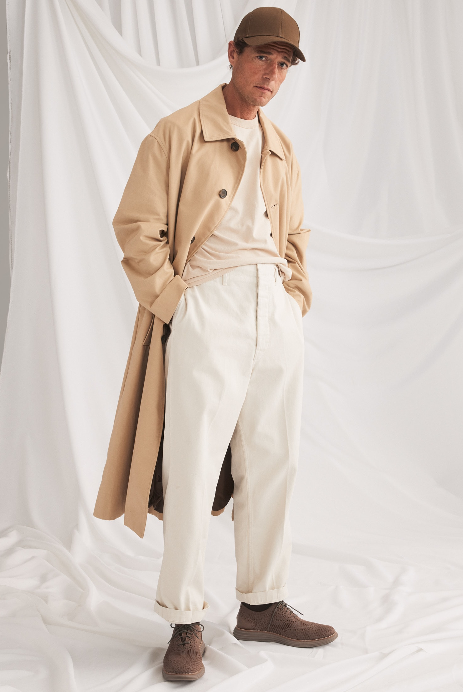 male model wearing oxfords and a long trench coat with a baseball cap