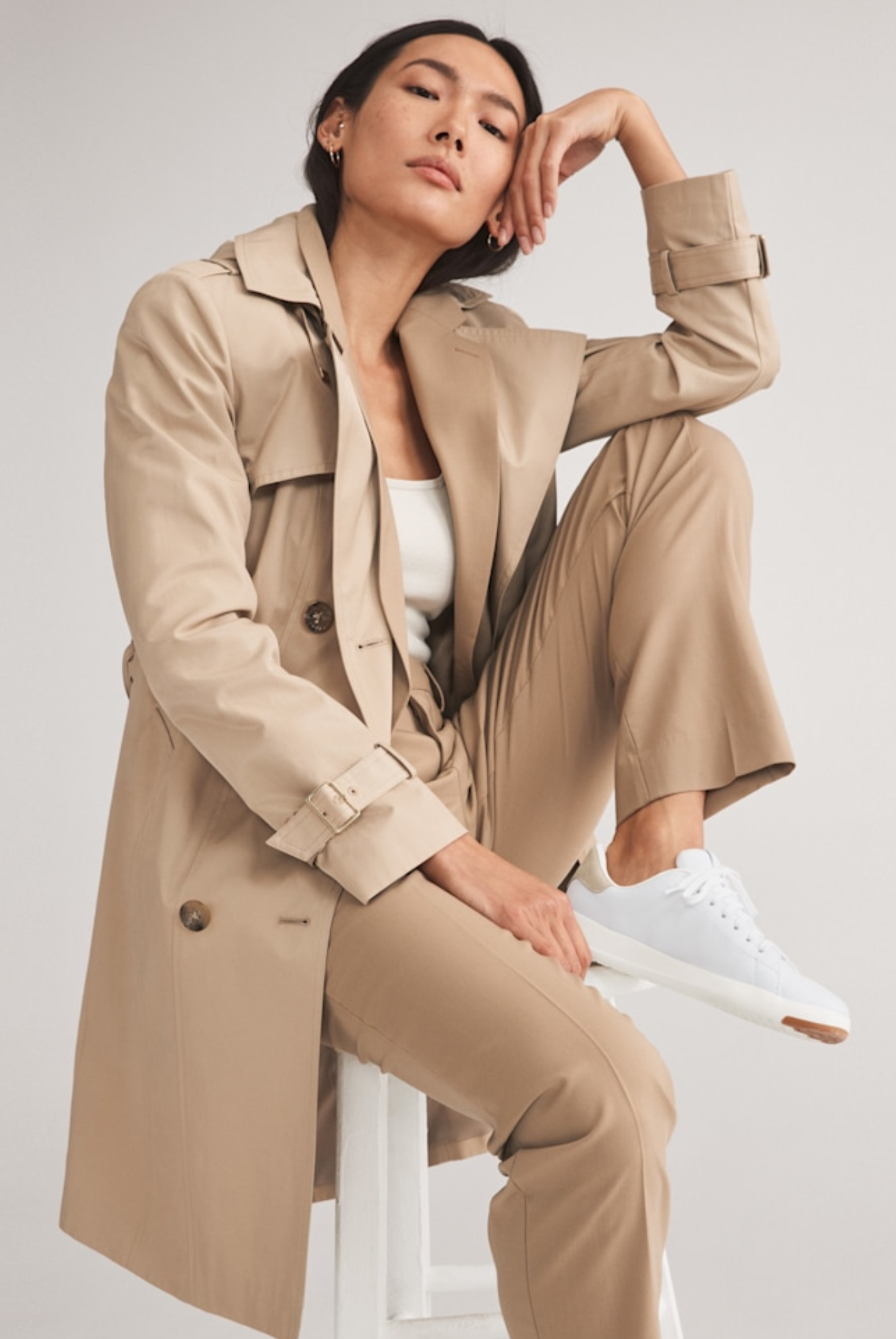 female model sitting on a stool while wearing a tan trench coat with matching pants and white sneakers